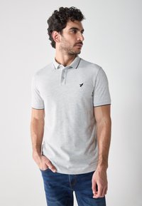 Polo majica od svijetlosive tkanine s suptilnom teksturom, kratkim rukavima i malim crnim logotipom na prsima. Ima kontrastni ovratnik.
