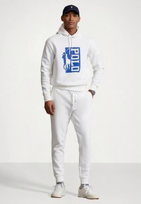 Balts džemperis ar zilu "POLO" logotipu un zirga grafiku, kopā ar saskaņotām baltām sporta biksēm. Aprīkots ar auklu jostasvietā un ribotas aproces.