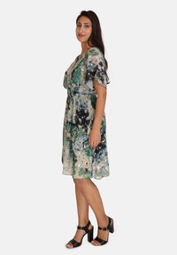 Bloemenjurk in tinten groen en blauw, lichtgewicht stof, elastische taille, fladdermouwen, gelaagd ontwerp, gecombineerd met zwarte blokhakken sandalen.