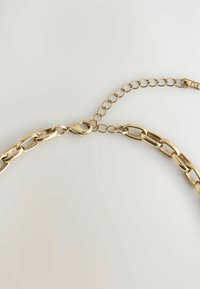 Goldfarbene Metallkette mit ovalen Gliedern, ausgestattet mit einem Karabinerhaken und einer verstellbaren Kettenverlängerung zur Anpassung der Passform. Glatte Textur.