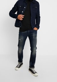Chaqueta de mezclilla oscura con botones en el frente y dos bolsillos en el pecho, combinada con una camisa negra, jeans desgastados y zapatillas negras.