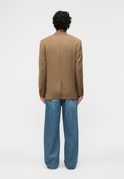 Άτομο με σγουρά μαλλιά, φορώντας καφέ σακάκι και φαρδιά μπλε τζιν, στέκεται με την πλάτη στραμμένη προς ένα απλό ανοιχτό φόντο.