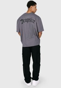 Graues Baumwoll-T-Shirt mit dem Aufdruck "Dropsize" in Schwarz auf dem Rücken. Kombiniert mit schwarzen Cargo-Hosen und weißen Sneakers.