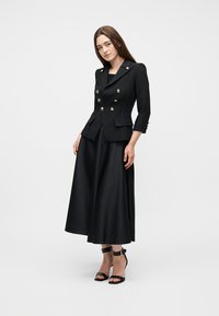 Γυναίκα που φοράει ένα μαύρο σακάκι με διπλό πέτο και χρυσά κουμπιά, μια μαύρη midi φούστα και μαύρα ψηλοτάκουνα παπούτσια με λουράκι στον αστράγαλο, στέκεται μπροστά σε λευκό φόντο.