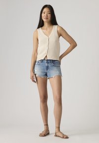 Νεαρή γυναίκα όρθια με το ένα χέρι στη μέση, φορώντας μπεζ αμάνικο τοπ με κουμπιά, φθαρμένα τζιν σορτς και καφέ φλατ σανδάλια.