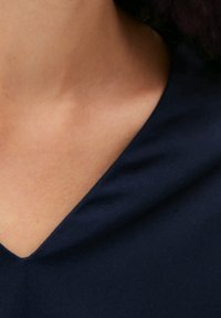 Primo piano del collo e della parte superiore del torace di una persona che indossa un indumento a V di colore blu scuro su uno sfondo neutro.