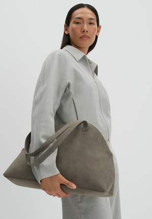 Frau in grauer Jacke mit Reißverschluss und Hose, die eine große taupefarbene Ledertasche vor einem schlichten hellen Hintergrund hält.