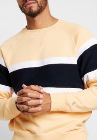 Man med beige, svart och vitrandig tröja med hopknäppta händer som visar tatuerade fingrar mot en enfärgad bakgrund.