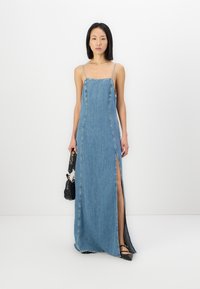 Denim maxi-kjole med tynne stropper, en rett silhuett, splitt i siden og råkant. Brukt med en liten svart veske og hælsko.