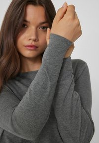 Šedý top s dlouhým rukávem vyrobený z měkké látky, s přiléhavými rukávy. Výstřih je široký a textura vypadá hladce a nenápadně.