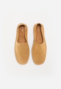 Bottines en daim beige à enfiler, présentant une semelle intermédiaire texturée et des coutures visibles le long des bords. Design minimaliste avec un bout arrondi.