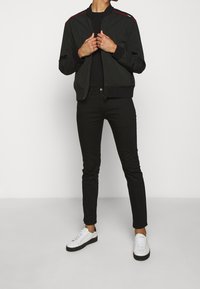 Zwarte bomberjack met rode accentstrepen, gecombineerd met zwarte skinny jeans en witte sneakers. De outfit heeft een gladde, strakke textuur.