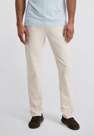 Mann trägt hellbeige gerade geschnittene Hose, hellblaues Hemd und dunkelbraune Loafers, steht vor einfachem Hintergrund.