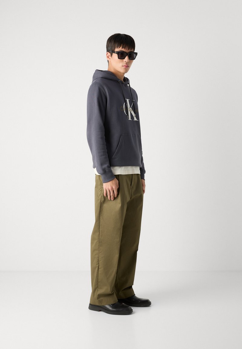 Sudadera gris con capucha y un logo, sobre una camiseta blanca. Pantalones anchos de color verde oliva, zapatos negros y gafas de sol rectangulares negras.