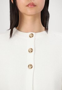 Cardigan blanc en tricot waffle avec un col rond et quatre boutons ronds de couleur marron clair sur le devant. Design simple et texturé.