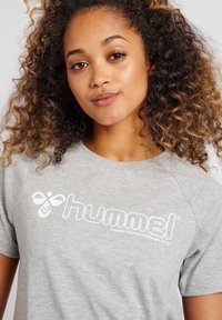 Grijze katoenen t-shirt met ronde hals en korte mouwen. Bevat een wit "hummel" logo en de slogan "VERANDER DE WERELD DOOR SPORT."