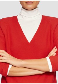 Rode gebreide trui met V-hals, over een witte turtleneck gedragen. Heeft raglan mouwen en contrasterende witte manchetten.