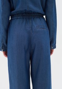 Denimu kombinezons ar elastīgu jostu, garām piedurknēm un aizmugurējam kabatām. Audums ir vidēji zils ar gludu tekstūru un redzamām dūrēm.