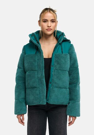 Frau trägt eine türkisfarbene Steppjacke mit Fleece-Textur und Kapuze, kombiniert mit schwarzen Hosen, vor einem schlichten hellen Hintergrund stehend.