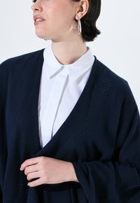 Marineblauer Cardigan mit weicher Textur, V-Ausschnitt und metallischen Akzenten, in Kombination mit einem weißen Hemd mit Kragen.