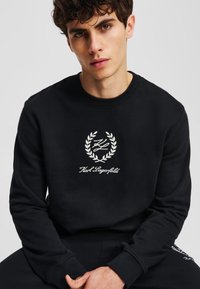 Fekete pulóver, kerek nyakkivágással, puha anyaggal és hímzett, babérkoszorú mintás logóval. Bordázott mandzsetták és szegély teszik teljessé a megjelenést.