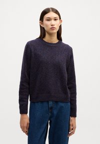 Dunkelvioletter Strickpullover mit Rundhals und langen Ärmeln, der ein strukturiertes Muster und einen gerippten Saum aufweist. Kombiniert mit hochgeschnittenen Jeans.