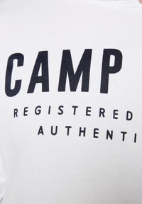 Witte katoenen shirt met gedurfde zwarte tekst die "CAMP" boven "REGISTERED" en "AUTHENTI" in kleinere letters weergeeft. Gladde textuur.