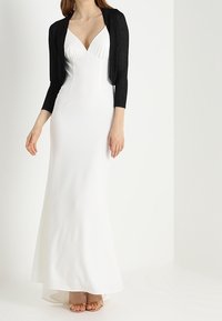Vestido de noche blanco con un corsé ajustado, escote en V profundo y falda fluida. Combinado con un cárdigan corto negro texturizado y tacones de tiras nude.