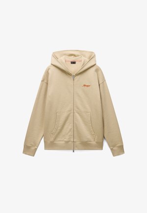 Bežová mikina na zip vyrobená z měkké tkaniny. Má velkou kapuci, přední kapsy a vyšívané logo na prsou. Jednoduchý, ležérní design.