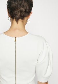 Bílý top s kulatým výstřihem, strukturovanou tkaninou a krátkými nafouklými rukávy. Vybaven zlatým zipem na zadní straně pro zapínání.
