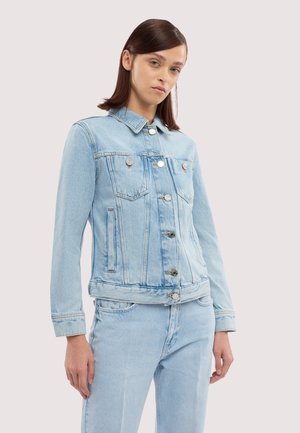 Giovane donna con capelli castani lisci che indossa una giacca di jeans azzurro chiaro e jeans abbinati su uno sfondo uniforme.