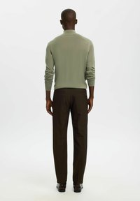 Homme debout, de dos, portant un haut à manches longues vert olive, un pantalon droit marron foncé et des chaussures habillées foncées, sur fond blanc.