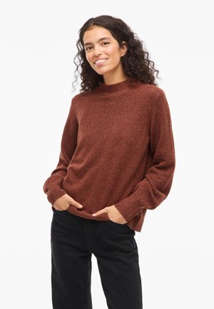 Mujer joven con cabello rizado, vistiendo un suéter de punto color óxido y pantalones negros, sonriendo con las manos en los bolsillos frente a un fondo blanco.