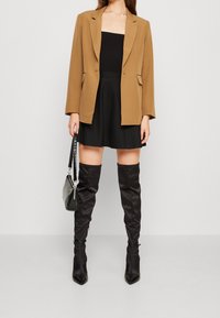 Hnědý blazer přes černý přiléhavý top, doplněný o rozšířenou černou sukni a černé kozačky nad kolena. Doplňky zahrnují malou černou kabelku.