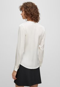 Langärmlige Bluse aus cremer Satin mit sanftem Glanz, mit abgerundetem Saum und Knopfmanschetten, kombiniert mit einem schwarzen Rock.