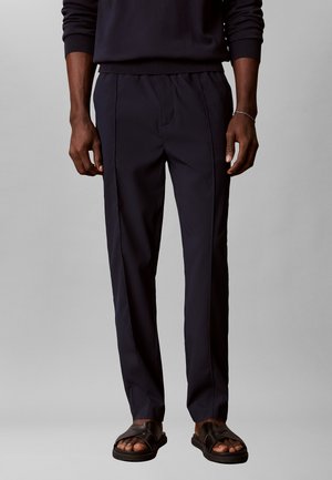 Mand iført sorte skræddersyede bukser, sort langærmet top og sorte lædersandaler med krydsede stropper på grå baggrund.