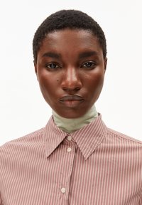 Camicia con bottoni di colore rosa con strisce bianche; presenta un colletto e chiusura con bottoni. Sotto è indossato un dolcevita verde chiaro, creando una texture stratificata.