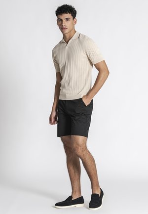 Giovane uomo in piedi, che indossa una polo a maniche corte beige in maglia, pantaloncini neri e scarpe slip-on nere con suole bianche, su uno sfondo chiaro uniforme.