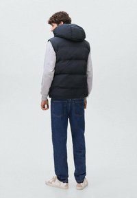 Črna pufasta brezrokavnik s kapuco, oblečen nad svetlo sivo majico s kapuco, v kombinaciji z modrimi kavbojkami in svetlimi športnimi copati. Vidne podrobnosti prešitja na brezrokavniku.