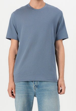 Mand iført en ensfarvet blå kortærmet t-shirt med rund hals og lyseblå denimjeans står foran en hvid baggrund.