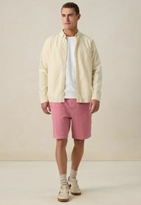 Chemise à rayures jaune clair sur un t-shirt blanc, associée à des shorts texturés rose et des baskets beige. Arrière-plan neutre.