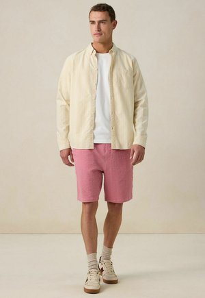 Chemise à rayures jaune clair sur un t-shirt blanc, associée à des shorts texturés rose et des baskets beige. Arrière-plan neutre.