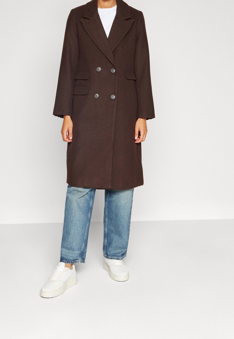 Personne portant un long manteau marron à double boutonnage, un jean bleu clair et des baskets blanches, debout devant un fond clair uni.