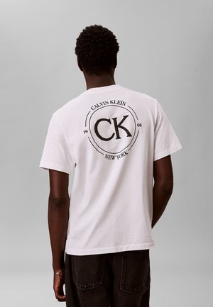 Persona con capelli corti e ricci che indossa una maglietta bianca Calvin Klein con logo e testo nero sul retro, in piedi davanti a uno sfondo grigio uniforme.