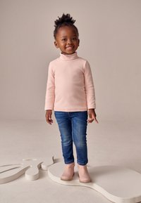 Lyserød rullekravebluse i let stof, blå jeans med let falmet effekt og lyserøde ankelstøvler, stående på en lys abstrakt form.