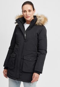 Parka noire matelassée avec capuche doublée en fourrure, fermeture éclair et accents de boutons. Comprend deux poches avant. Texture douce, design chaud.