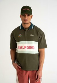Grøn polo t-shirt med horisontale hvide og lyseblå striber, prydet med "BERLIN SOHO" i pink tekst og farverige broderede detaljer.