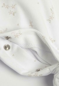 Tessuto di cotone bianco con piccoli motivo floreali in rosa chiaro e oro. Presenta una chiusura a scatto su un lato e una texture liscia.