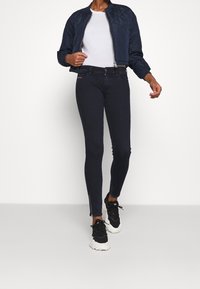 Marinblå quiltad bomberjacka, vit t-shirt, mörka tajta jeans med sidodragkedjor, svartvita sneakers. Avslappnad outfit visad.