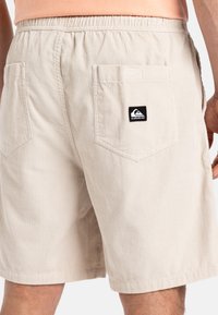 Pantalones cortos de pana beige con una cinturilla elástica, dos bolsillos traseros y un parche del logo negro en el bolsillo derecho.